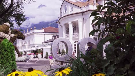 people are walking along the spa promenade whilst others are taking pictures and selfies in the city center of meran, just opposite the kurhaus