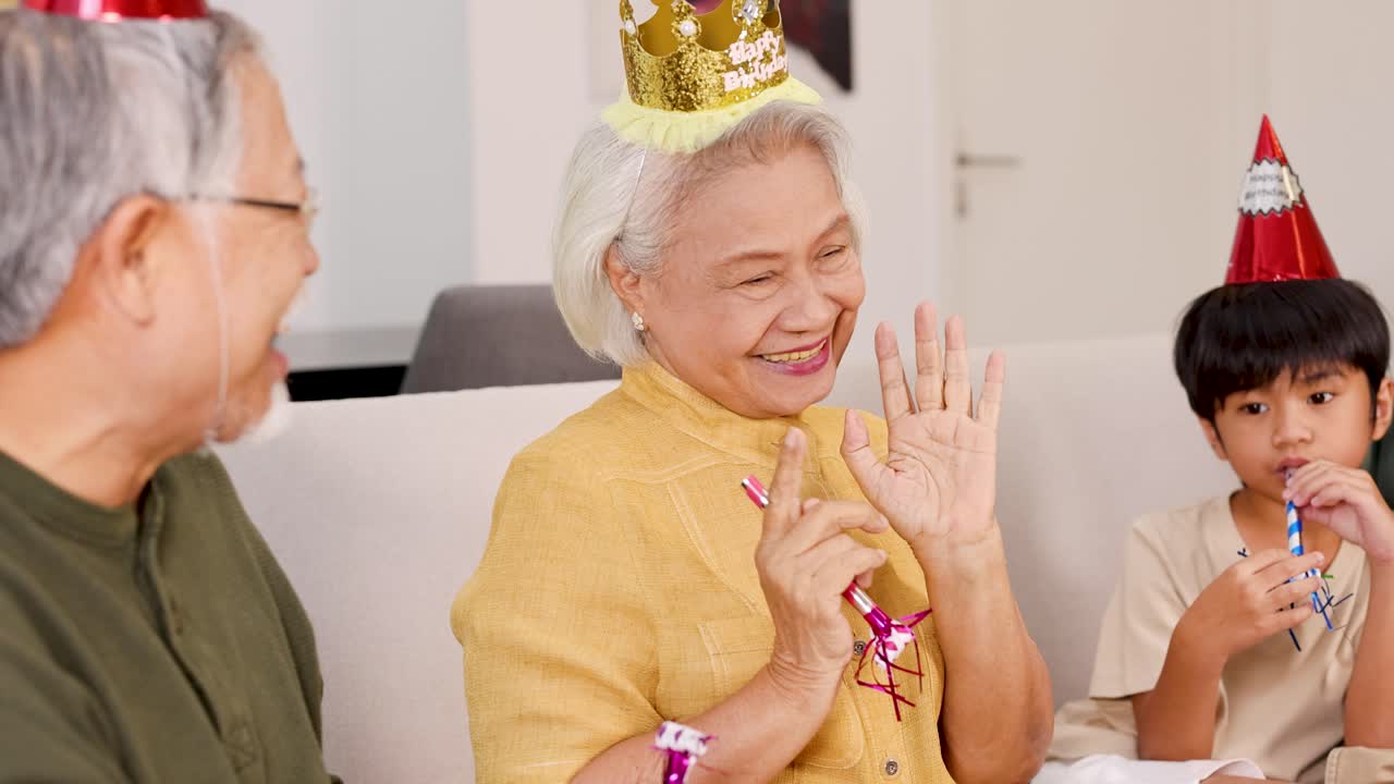 Elderly couple and child celebrate a birthday with laughter, wearing festive hats in a warmly lit room