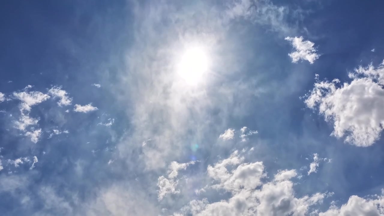4K UHD timelapse: bright radiant sun dominates expansive blue skyscape. Shifting misty cloud cover and scattered lower cumulus formations capture atmospheric diffusion and high-altitude weather.