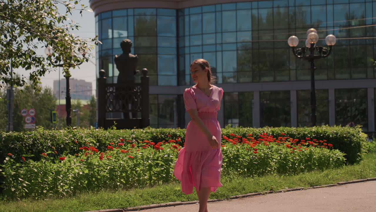 Mulher branca vestindo um vestido rosa, passeando por um parque urbano ao lado de um edifício de vidro, girando perto de canteiros de flores, posando perto de cercas vivas, prendendo o cabelo, luz solar suave filtrando-se pelas folhas, clima romântico e relaxante
