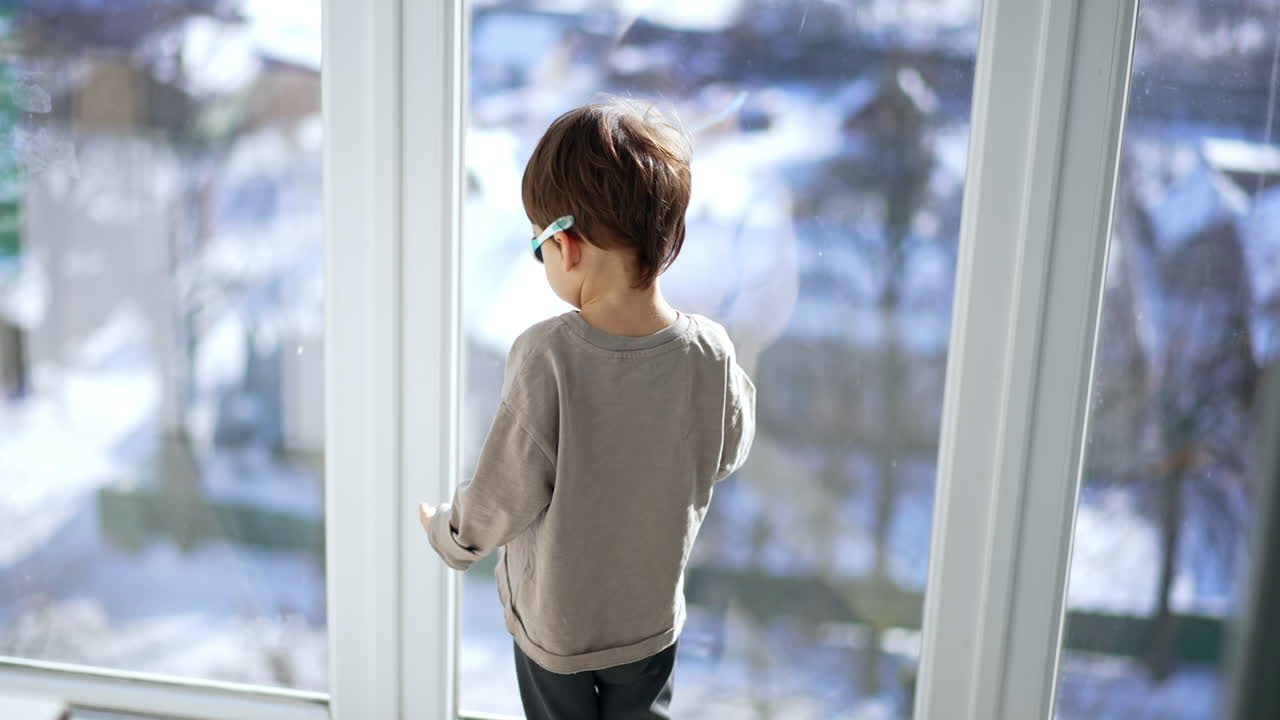 Rear view of a toddler standing near the panoramic window. Kid in sunglasses on balcony. Child turns around and goes to the room.