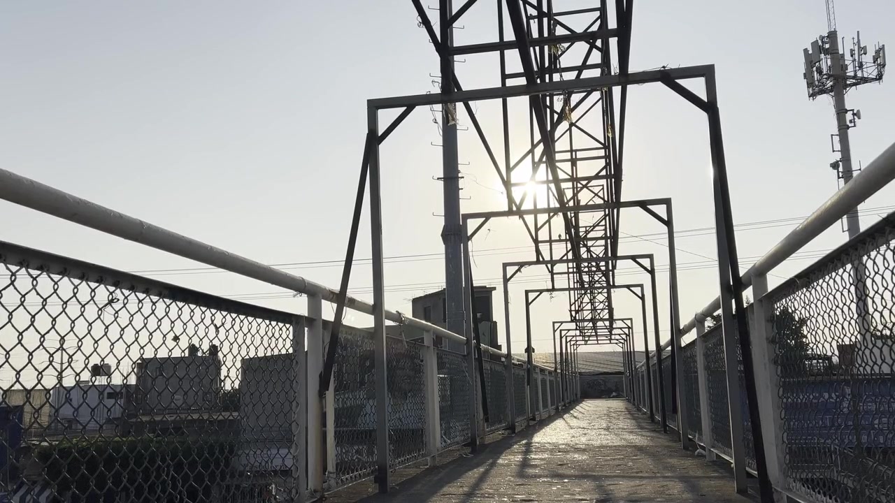 A perspective view of an industrial pedestrian bridge with metal beams and chain-link fencing, illuminated by the setting sun in an urban setting.