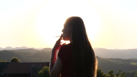 Young-woman-standing-outside-smoking-cigarette-and-enjoying-the-view-of-landscape-with-sunset