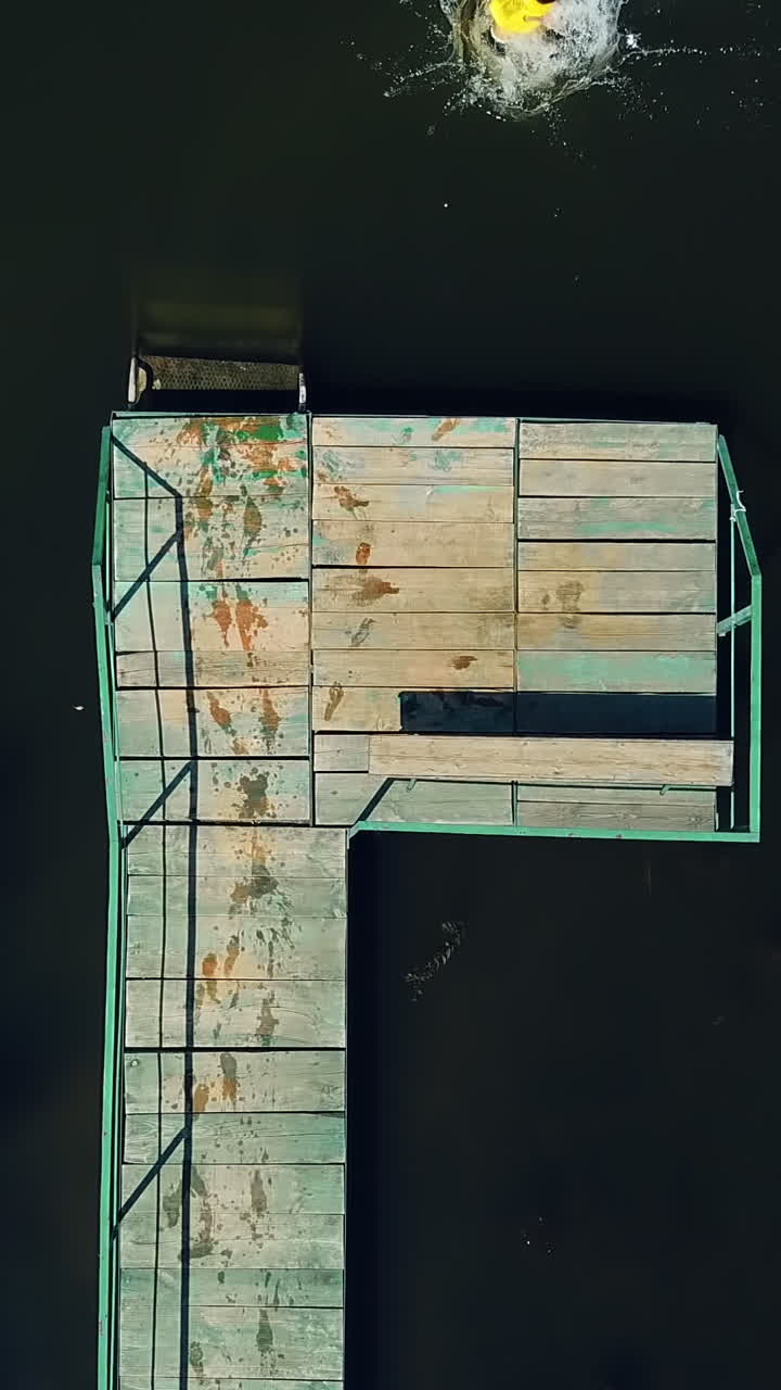 Little boy walking on the long wooden bridge and jumping into the water in a sunny day. Kid wearing waistcoat having a good time in the river on his summer holidays. Aerial view Vertical video