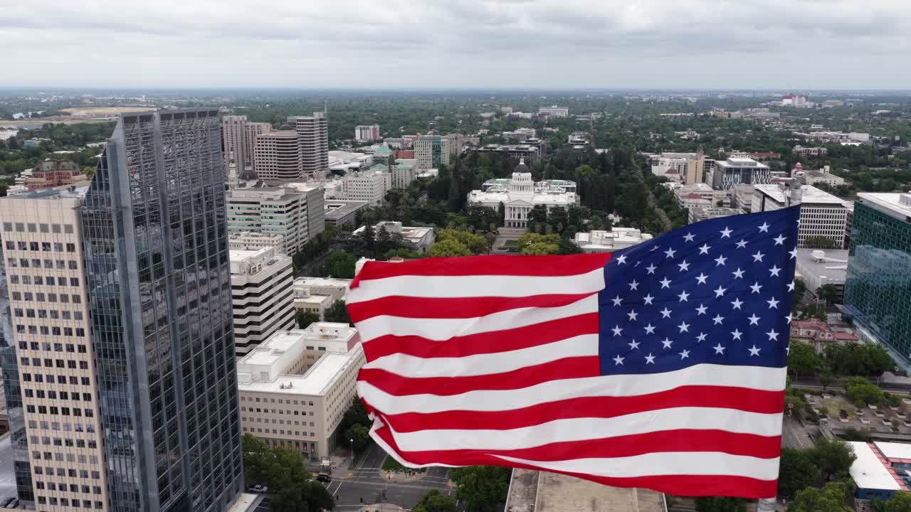 Rivelazione aerea della bandiera sul centro di Sacramento con una grande bandiera americana che sventola sopra lo skyline della città e il Campidoglio dello Stato della California sullo sfondo. Scena patriottica di un punto di riferimento urbano sotto la luce diurna nuvolosa. Ampia.