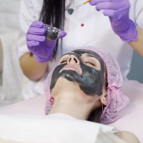 an experienced cosmetologist carefully applies a black cleansing mask to a woman's face in a beauty salon. close-up. industry of beauty.