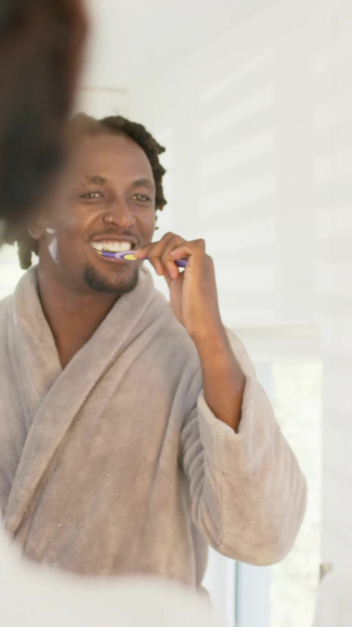 Vertical video: Facing mirror, man brushing teeth in bathroom in robe, using toothbrush for hygiene