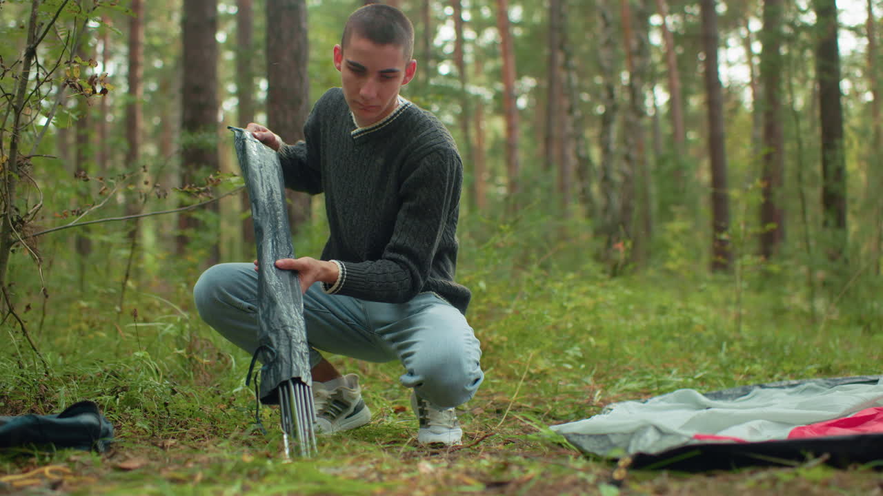 Young man squats on forest ground holding tent pole bag as he begins to unpack and adjust poles for setting up tent, surrounded by green foliage, trees, and scattered gear during outdoor camping