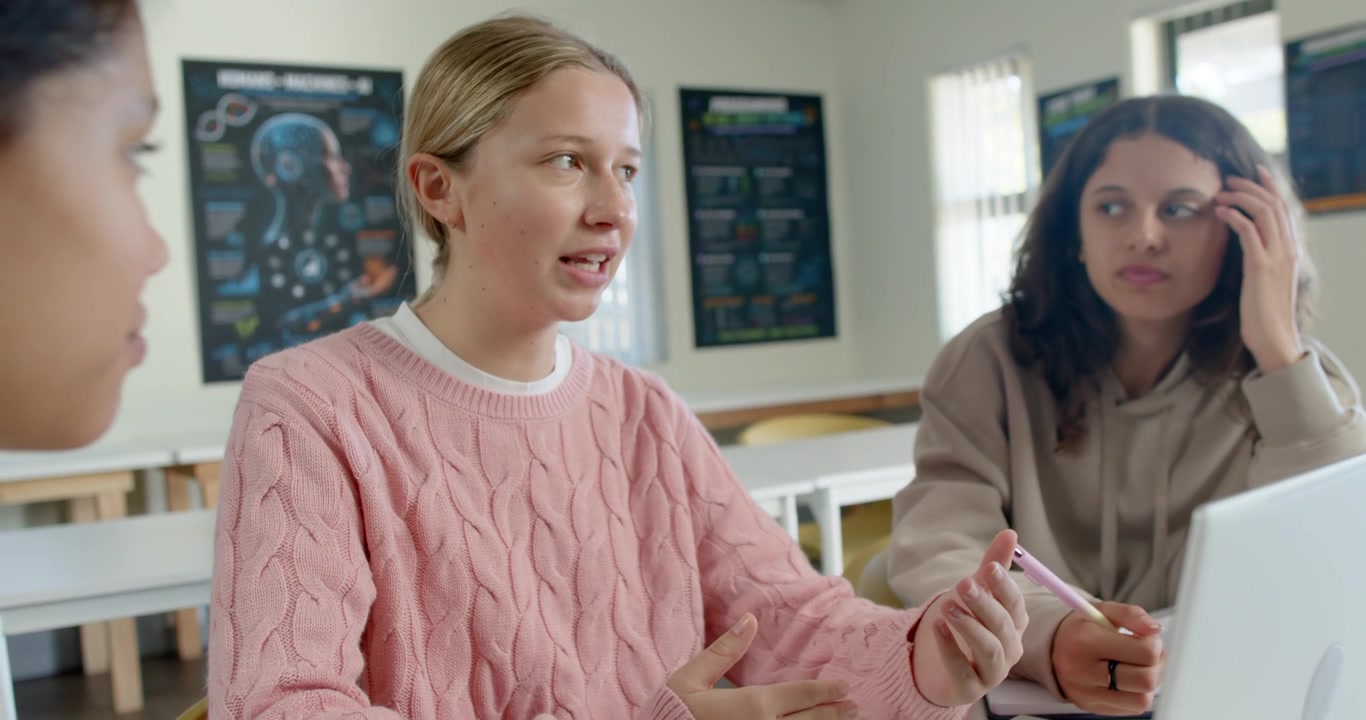 Teen girls in class being prompted blonde in pink sweater speaking while brunette noting on laptop