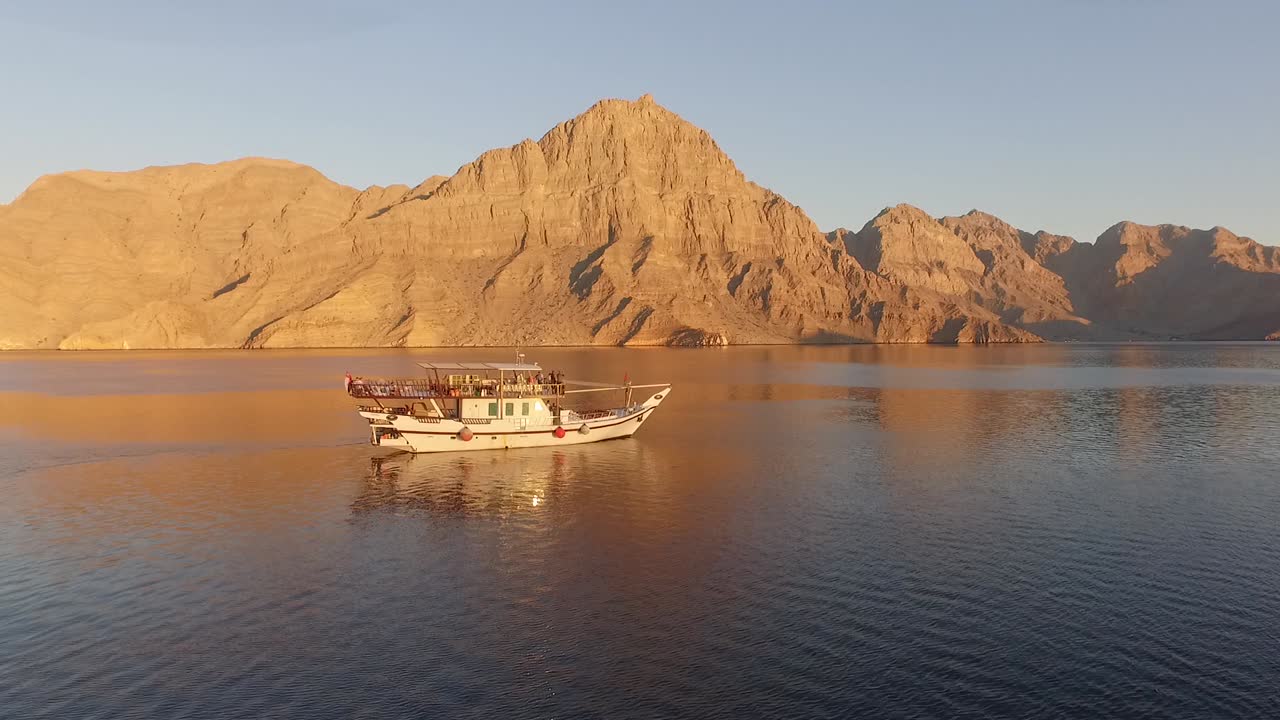 Drone che sorvola una barca tradizionale nello Stretto di Hormuz, Oman, vicino all'Iran, con le spettacolari montagne del Musandam riflesse nel mare calmo, lungo una delle rotte marittime più strategiche del mondo