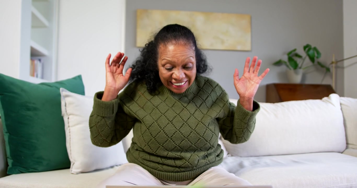 Senior African woman reading email on laptop on sofa smiling, clapping, leaning toward screen