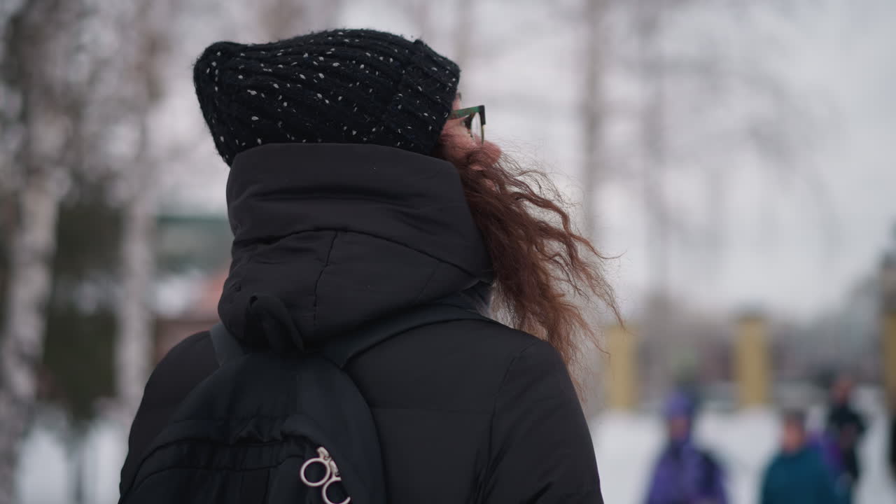 검은색 코트, 모자, 안경, 백팩을 착용하고 눈 덮인 도시 공원 겨울에 서 있는 곱슬머리 여성, 추운 분위기가 보이고 배경은 흐릿하며 사람들이 걷는 얼어붙은 환경