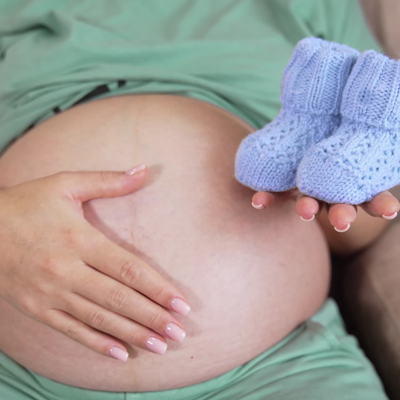 Donna incinta seduta sul divano che accarezza la sua grande pancia. Sta allungando la mano destra con dei piccoli calzini da neonato. Primo piano di scarpine da neonato blu.
