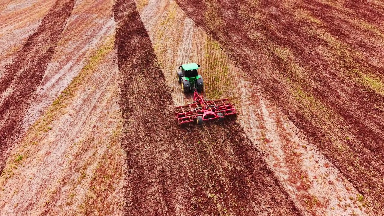 Una macchina verde contrasta nettamente con il terreno dalla texture rosso-ocra, con solchi diagonali che riempiono l'intera inquadratura. Trattore che traina un erpice a dischi rosso attraverso i campi agricoli bulgari.