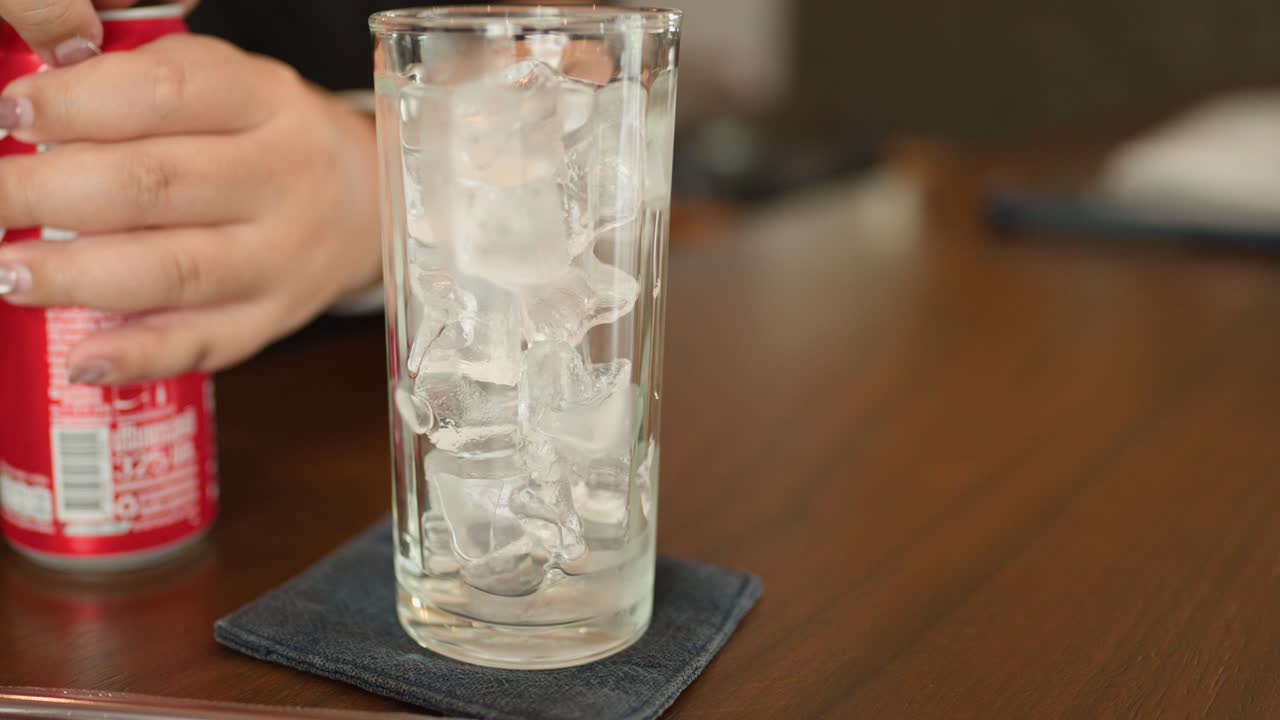 Hand pours cola from can into ice-filled glass on wooden table, natural lighting