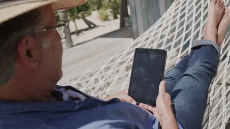 senior caucasian man in sunhat sitting in hammock using tablet by beach bar, in slow motion