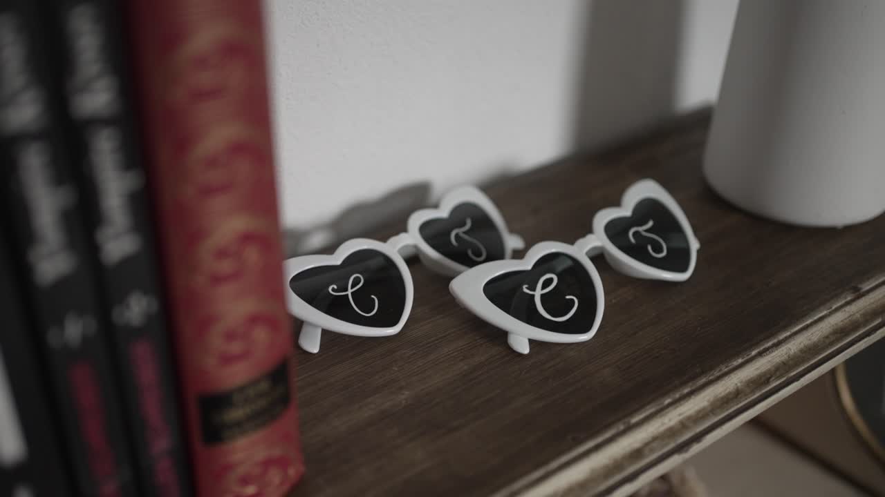 white heart shaped sunglasses with cursive initials resting on wooden shelf next to books