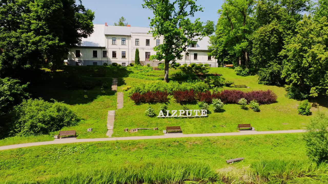 Green park with Aizpute sign and historic building on sunny countryside day