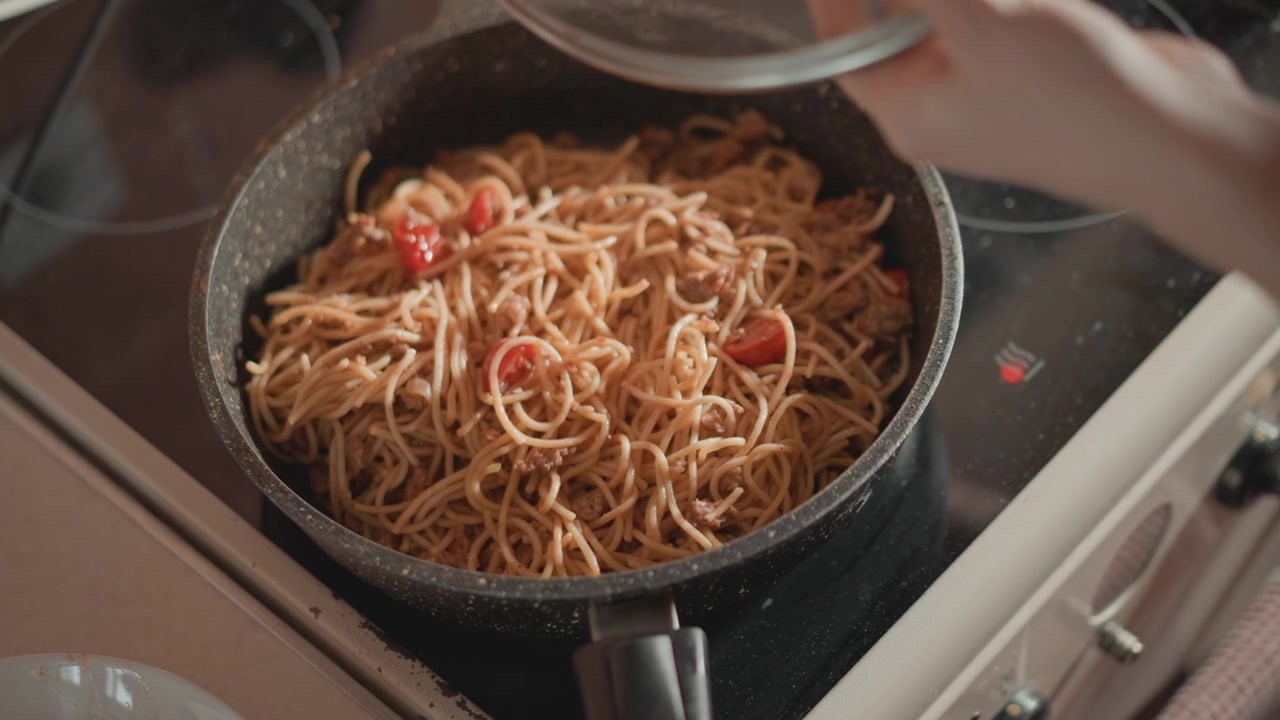 Vista ravvicinata dall'alto di spaghetti che cuociono in una padella scura su un fornello moderno, una mano posa un coperchio di vetro sulla pasta mentre pezzi di pomodoro, olio e vapore creano un pasto casalingo caldo a base di spaghetti.