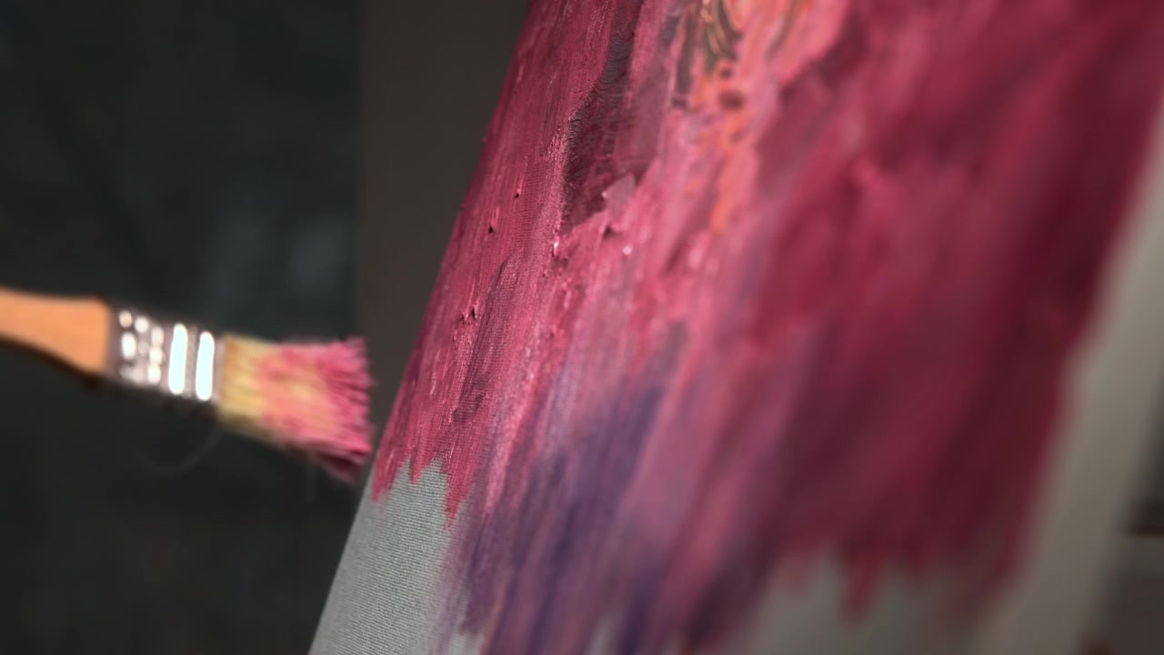 Primo piano di una mano che applica pittura rosa e viola con un pennello su tela texturizzata, creando pennellate astratte ed espressive su uno sfondo scuro.
