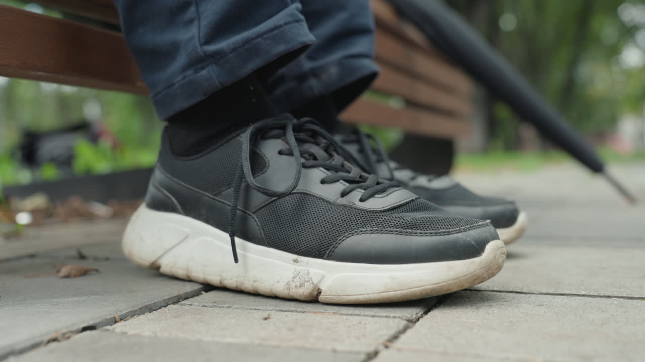 Sneaker set fra siden på fortov ved bænk, løber hviler sig mellem omgange, strikket sort sko med hvid sål, snavset slidbane, afslappet positur, grønt område i parken, antydning af bevægelse, fitnessrutine, restitution