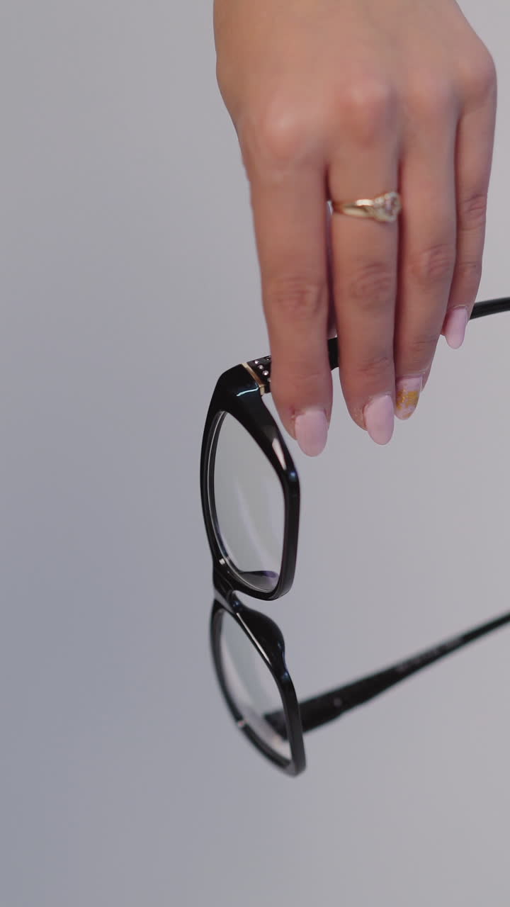 female hand is holding model of eyeglasses with black rim on a gray background. Vertical video