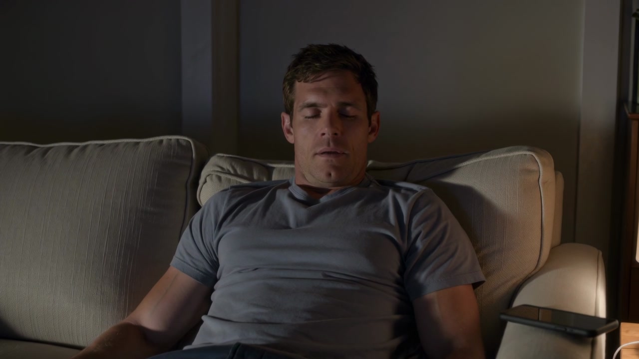 Waking to lamp light, man in gray T-shirt opening eyes, sitting on living-room sofa beside tablet