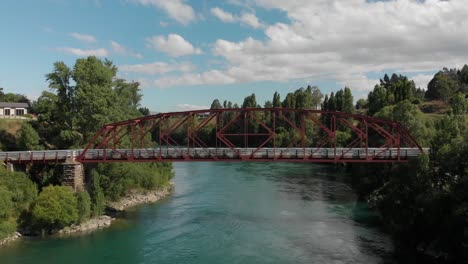 Aerial---Red-bridge-over-beautiful-blue-river-in-Clyde,-Central-Otago,-New-Zealand-with-cyclists-biking-over