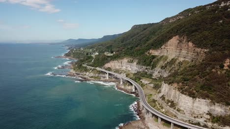 aerial: high elevation drone shot tracking along side a sea cliff bridge as cars drive across during a sunny day in new south wales, australia