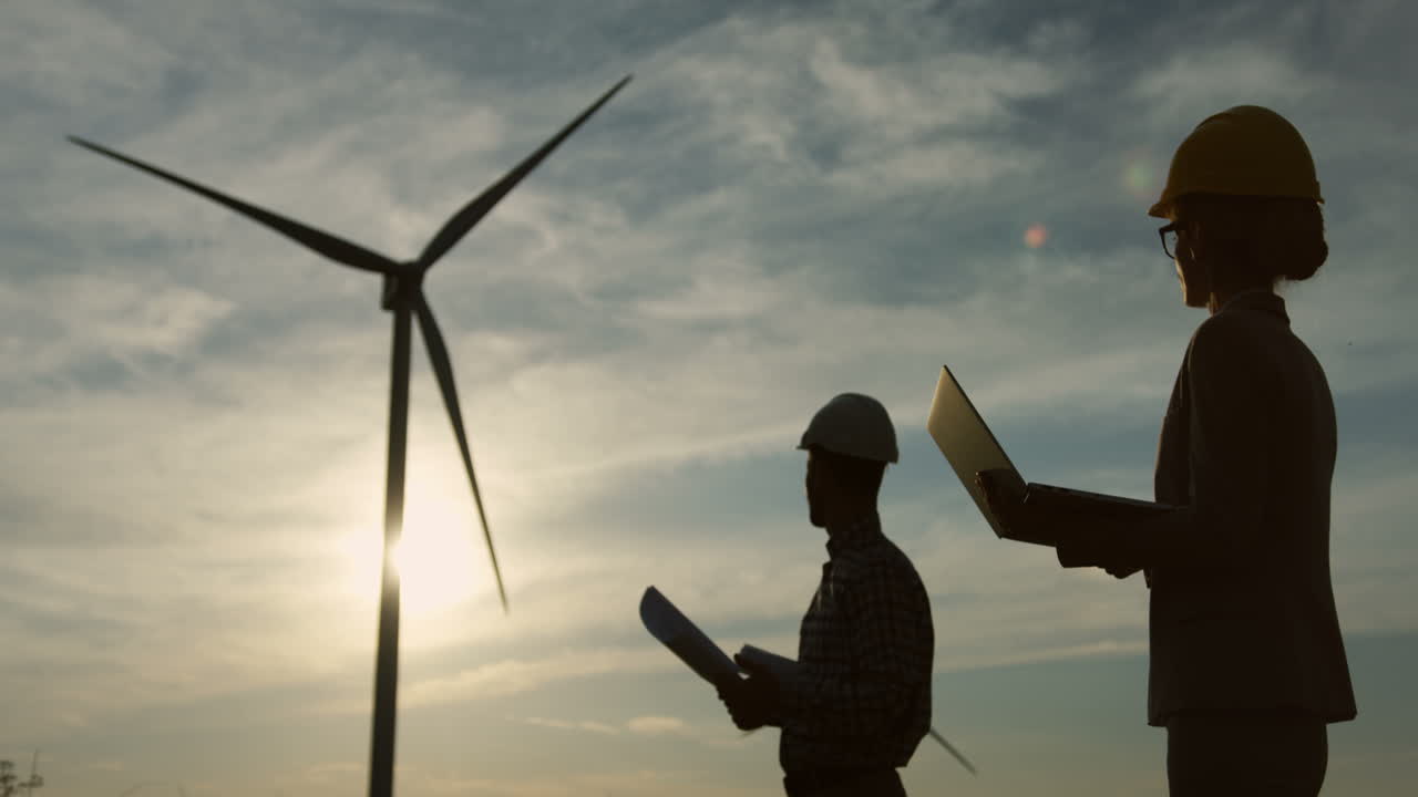 헬멧을 쓰고 재생 에너지 풍력 발전소에서 문서나 청사진을 읽는 백인 여성과 남성 엔지니어의 실루엣