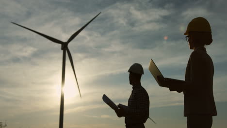 siluetas de una mujer caucásica y un hombre ingenieros usando un casco y leyendo algunos documentos o planos en la estación eólica de energía renovable