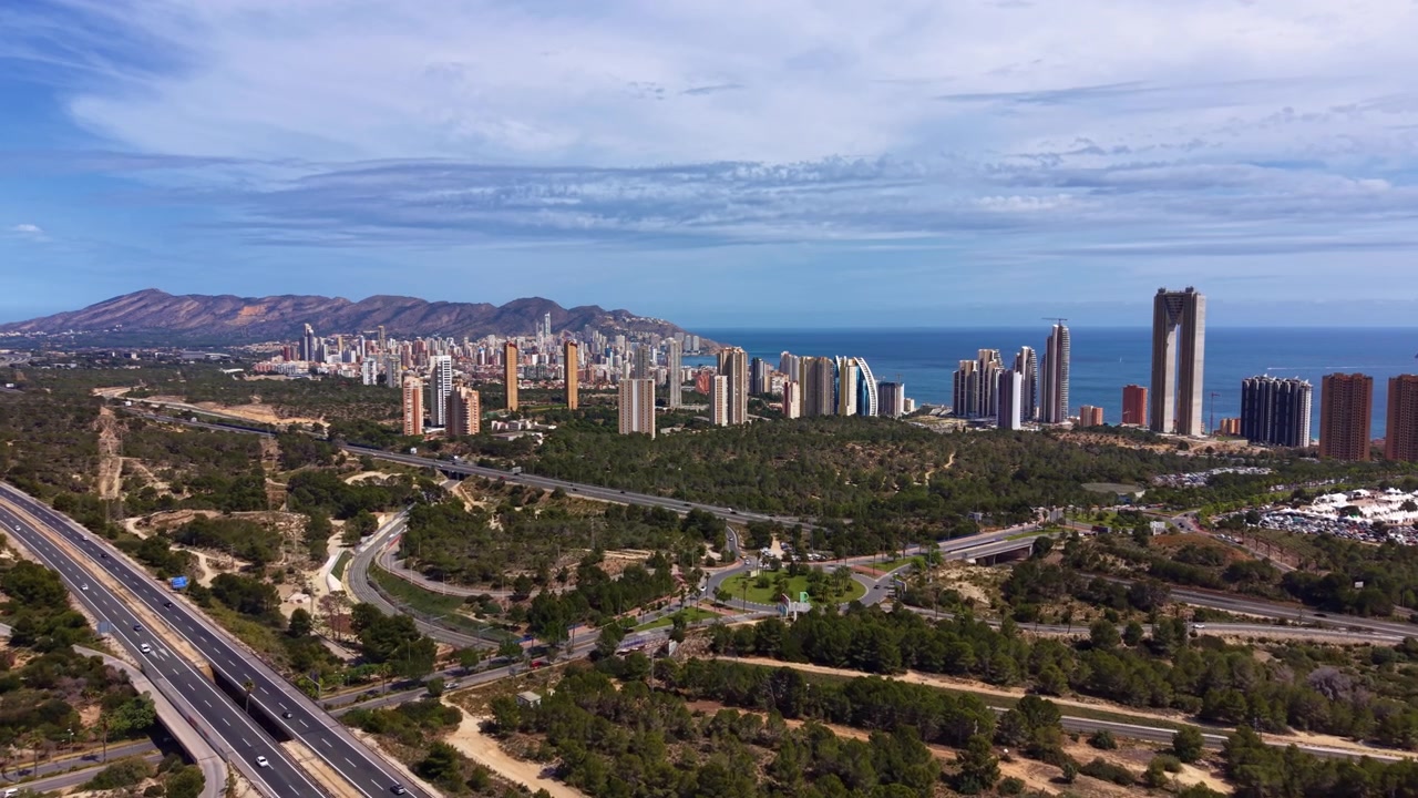 Veduta panoramica di Benidorm, Spagna, che mostra grattacieli lungo la costa mediterranea, un'autostrada trafficata, una foresta verde e montagne sotto un cielo blu.