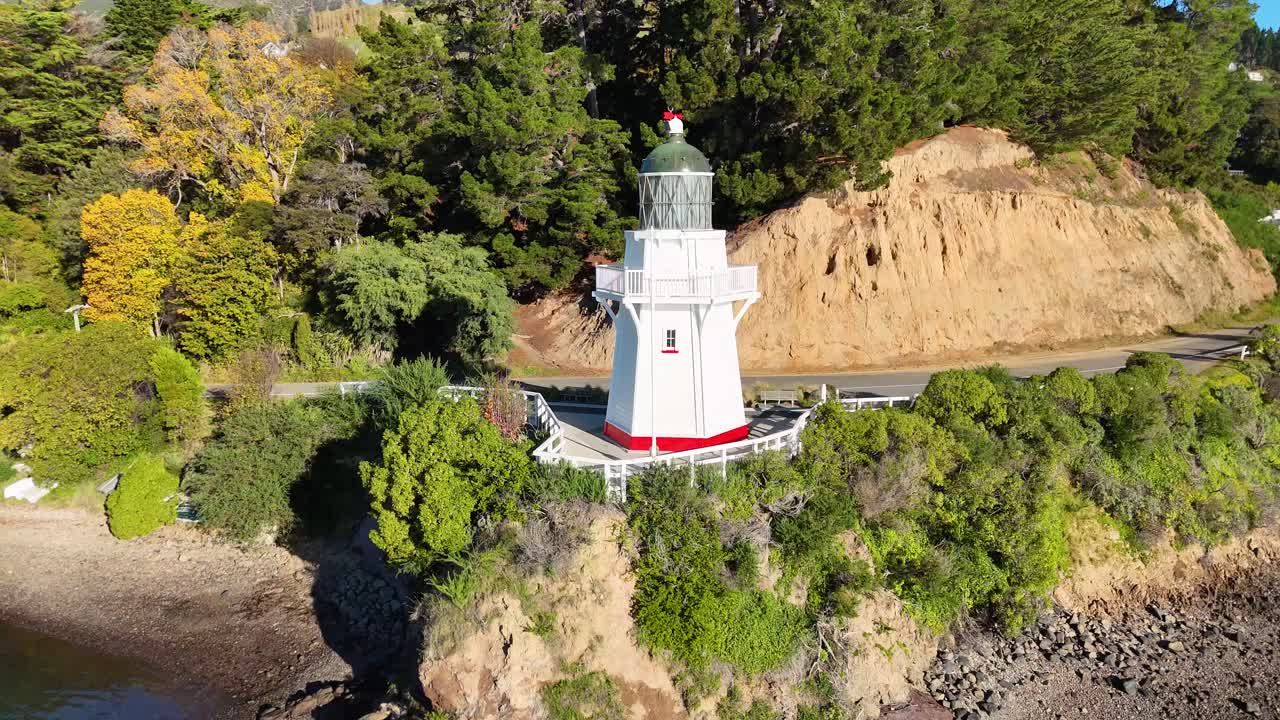 Drone footage captures Akaroa Lighthouse amidst lush greenery and calm waters in bright daylight