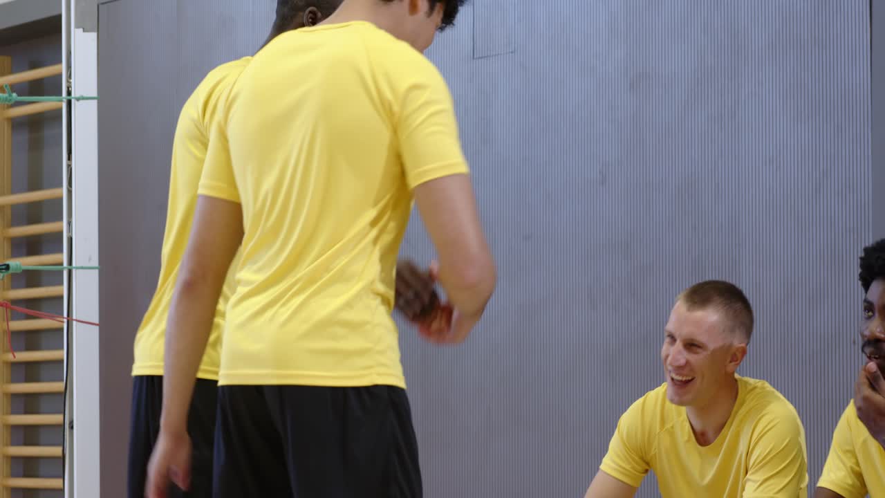 Squadra maschile eterogenea in giallo saluta il giocatore che arriva, il quale, con il pallone da pallavolo, guarda il telefono in palestra.