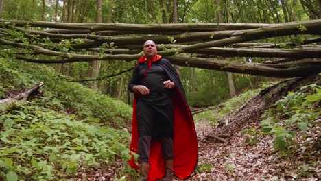 portrait of a man dressed up in black and red cape taking off his hood while walking on a trail in the forest