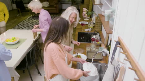 vista superior de una mujer lavando los platos de la cena familiar en el fregadero de la cocina mientras dos mujeres maduras quitan los platos de la mesa