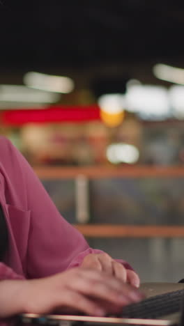 dama de vestido rosa sentada en un centro comercial, centrada en trabajar con su computadora portátil, el fondo está ligeramente borroso con personas moviéndose, con efectos de luz bokeh y otros objetos poco claros