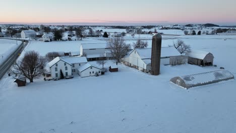 Vista-Aérea-De-Una-Granja-Cubierta-De-Nieve-Al-Atardecer-Con-Varios-Edificios-Y-Un-Silo.