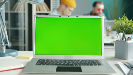 chromakey portátil de pantalla verde en el escritorio con empleados moviéndose en segundo plano en la oficina