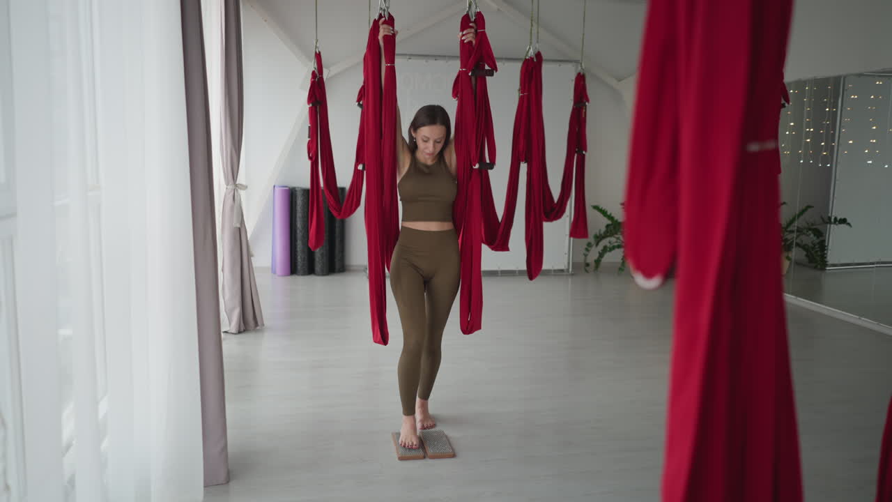 Flexible yoga instructor grips red silk hammock overhead while balancing legs atop two wooden nail boards in spacious studio performing acupressure and balance exercise for strength and inner calm