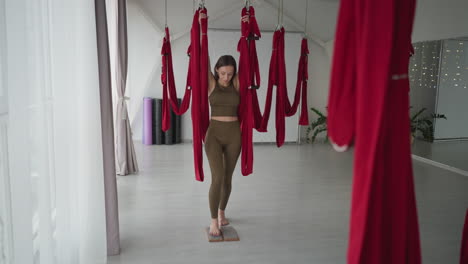 flexible yoga instructor grips red silk hammock overhead while balancing legs atop two wooden nail boards in spacious studio performing acupressure and balance exercise for strength and inner calm