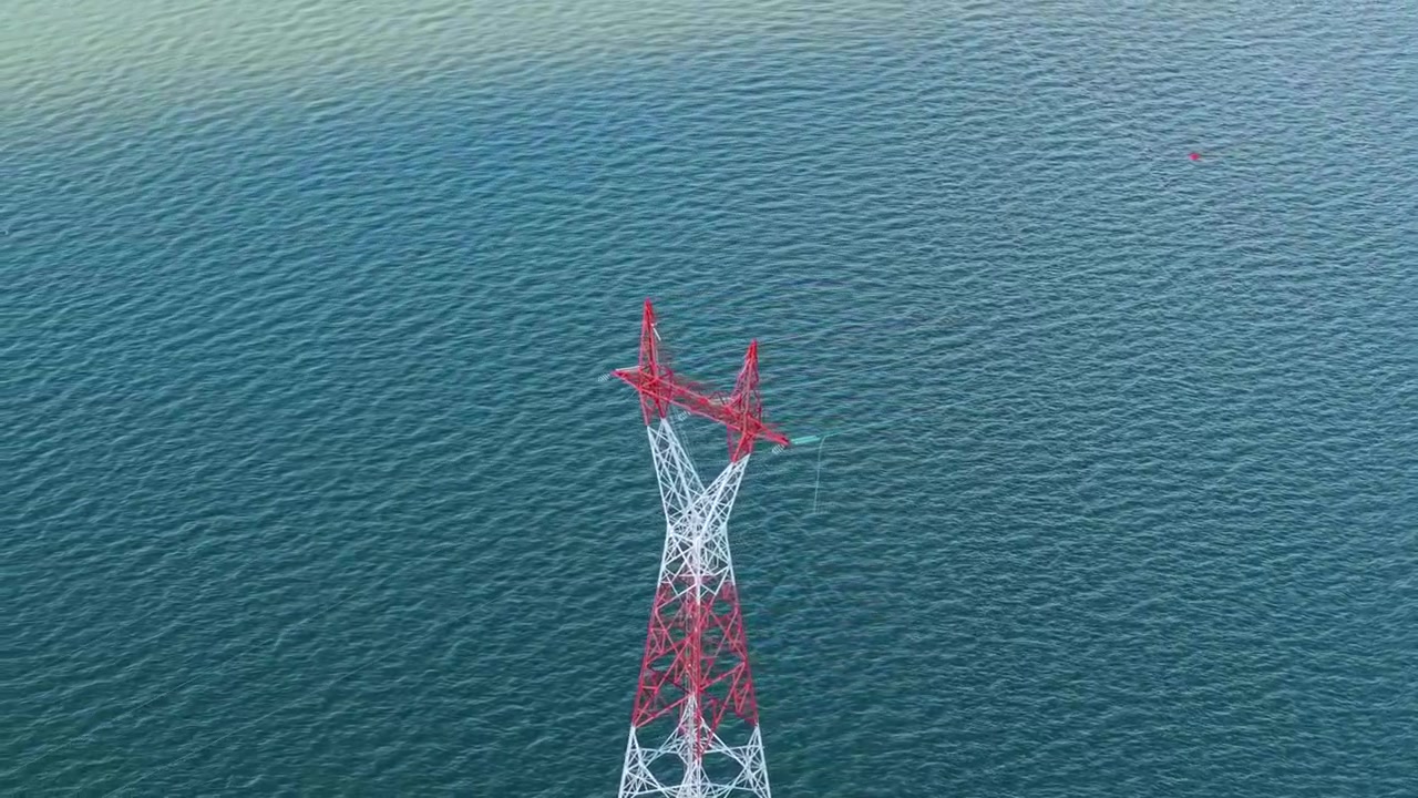 High voltage transmission tower stands directly in open water, red and white pylon structure supporting power line crossing over lake surface, drone orbit.