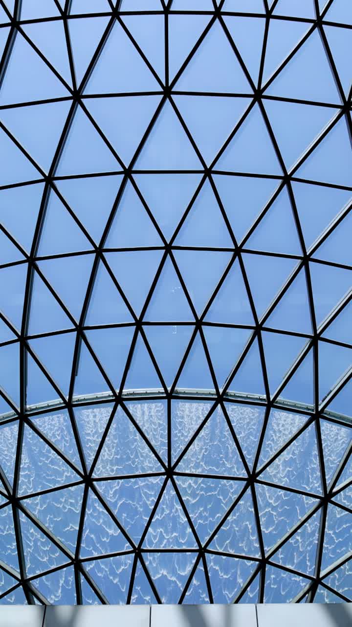 La telecamera si inclina verso l'alto sotto una grande cupola di vetro geometrica, rivelando una cascata interna circolare e la luminosa luce naturale del giorno. Ambiente architettonico moderno, movimento fluido della telecamera.