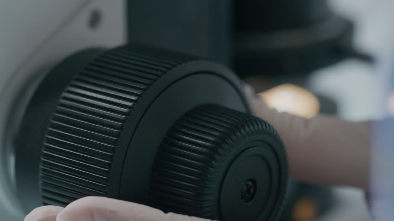Gloved Hand of Scientist Adjusting Microscope Focus Knob in Laboratory