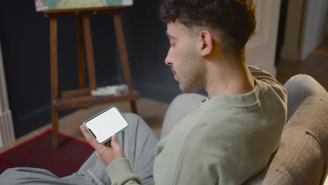 Vista-Sobre-El-Hombro-De-Un-Joven-Feliz-Sentado-En-El-Sofá-Y-Viendo-Algo-En-El-Teléfono-Inteligente