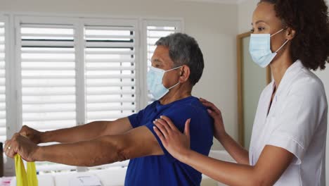 Physiotherapeutin-Mit-Gemischter-Abstammung-Trägt-Eine-Maske-Und-Hilft-Senioren-Beim-Training-Mit-Einem-Übungsband