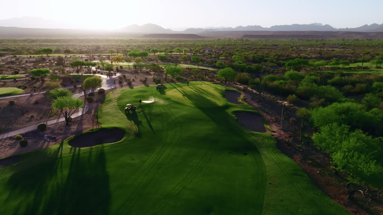 Push-in drone shot of a desert golf course with bunker, flagstick, green fairway, and cacti backed by rugged mountains as greenskeepers tend and water the green