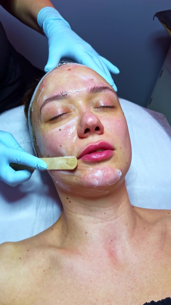 Una donna riceve un trattamento viso in un salone di bellezza di giorno, indossando una fascia per capelli e guanti blu.