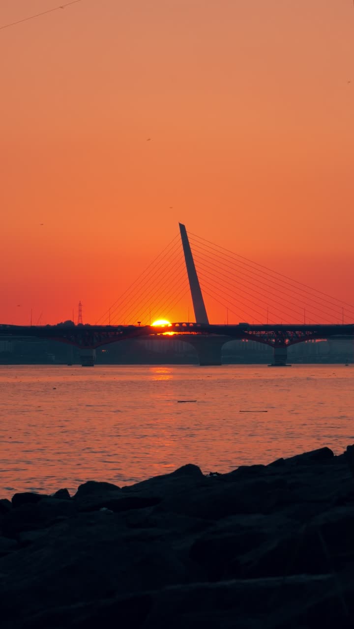Serene sunset over the Han River in Seoul, with the modern bridge silhouetted against the vibrant orange sky and the water shimmering below