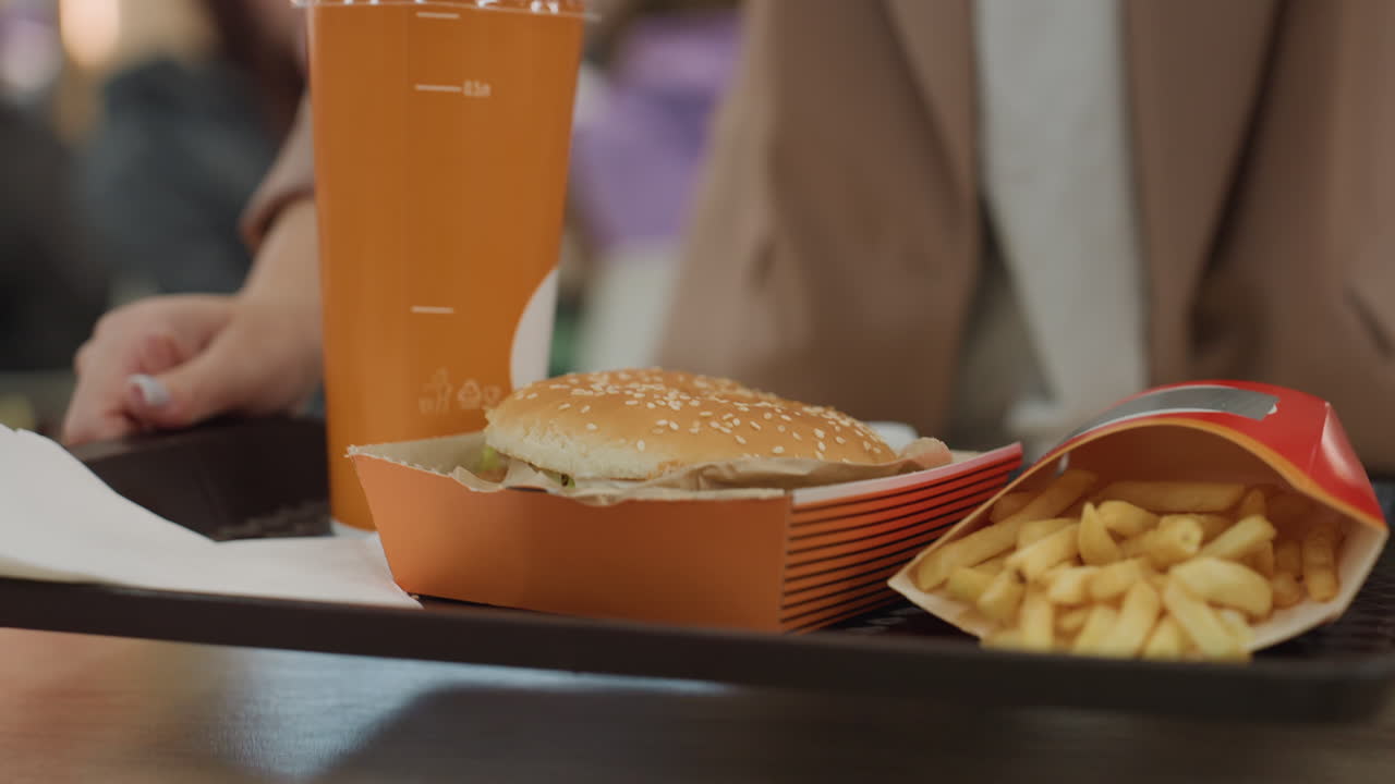 Mujer caucásica que lleva una bandeja de comida rápida con hamburguesa, patatas fritas y bebida en un concurrido patio de comidas de un centro comercial, abrigo beige, primer plano de las manos y las uñas cuidadas, escena neutral de viajeros con bullicio de fondo y mostrador